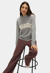 Grijze sweatshirt met "NORDIC" in crème tekst, geribbelde halslijn en boorden, gecombineerd met donker bordeaux brede broek, zittend op een kruk.