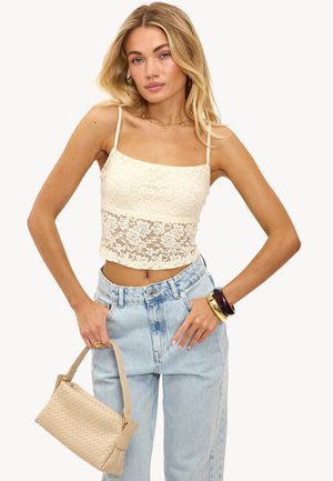 Top court en dentelle crème avec des bretelles fines, associé à un jean en denim bleu clair taille haute. Les accessoires incluent un sac à main en beige tressé et des bracelets.
