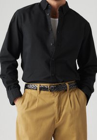 Schwarzes Hemd mit Knopfleiste, weicher Textur und klassischem Kragen, kombiniert mit beigen Hosen und einem geflochtenen schwarzen Ledergürtel.