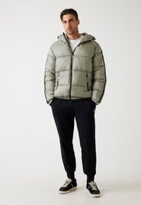 Chaqueta acolchada verde claro con capucha, cierre frontal y acentos laterales negros, combinada con pantalones de chándal negros y zapatillas negras.