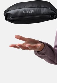 Hand mit nach oben geöffnetem Handflächen unter einem schwarzen Reißverschlusspouch mit der Aufschrift "STOW AWAY" vor einem schlichten hellen Hintergrund.
