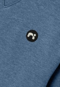 Tejido de algodón azul con una textura suave, que presenta un logotipo circular bordado en negro con un diseño blanco. Cuello entallado.