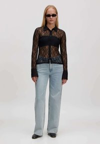 Vrouw in een zwarte kanten overhemd met knopen, lichtblauwe high-waisted jeans, zwarte vierkante schoenen en slanke zonnebril, staande tegen een effen achtergrond.