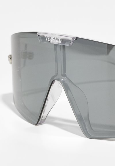 Gray oversized sunglasses with a geometric shape, clear accents, and a visible Versace logo at the top. Translucent frame.