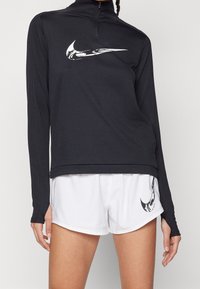 Czarna bluzka z długim rękawem, z wysokim kołnierzem i pół-zamkiem, z wyraźnym białym logo Nike; w połączeniu z białymi szortami z logo.