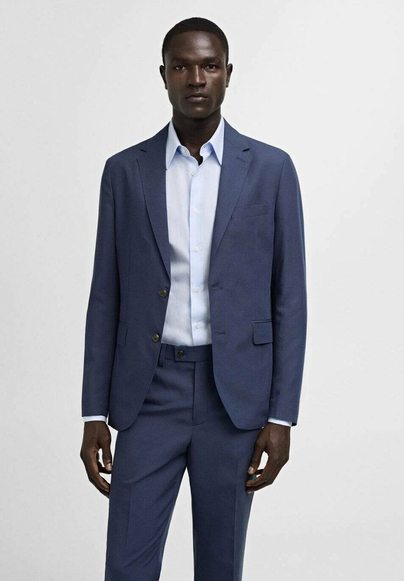 Marineblaues Anzug mit einer taillierten Jacke und passender Hose, kombiniert mit einem hellblauen Hemd. Glattes Material und maßgeschneiderter Schnitt.