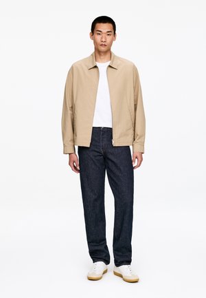 Mężczyzna stoi ubrany w beżową bluzę na zamek, białą koszulkę, ciemnoniebieskie dżinsy i białe trampki z brązową podeszwą na białym tle.