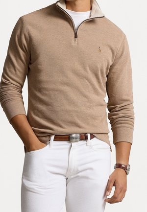 Man draagt een beige trui met een rits tot aan de hals over een wit overhemd, witte broek met een bruine riem en een bruine horloge, handen ontspannen langs het lichaam.