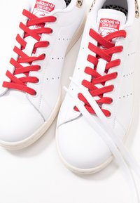 Weiße Leder Adidas Stan Smith Sneaker mit roten Schnürsenkeln, durchbrochenen Seitendetails und einer beigefarbenen Gummisohle. Zusätzlich sind weiße Schnürsenkel enthalten.
