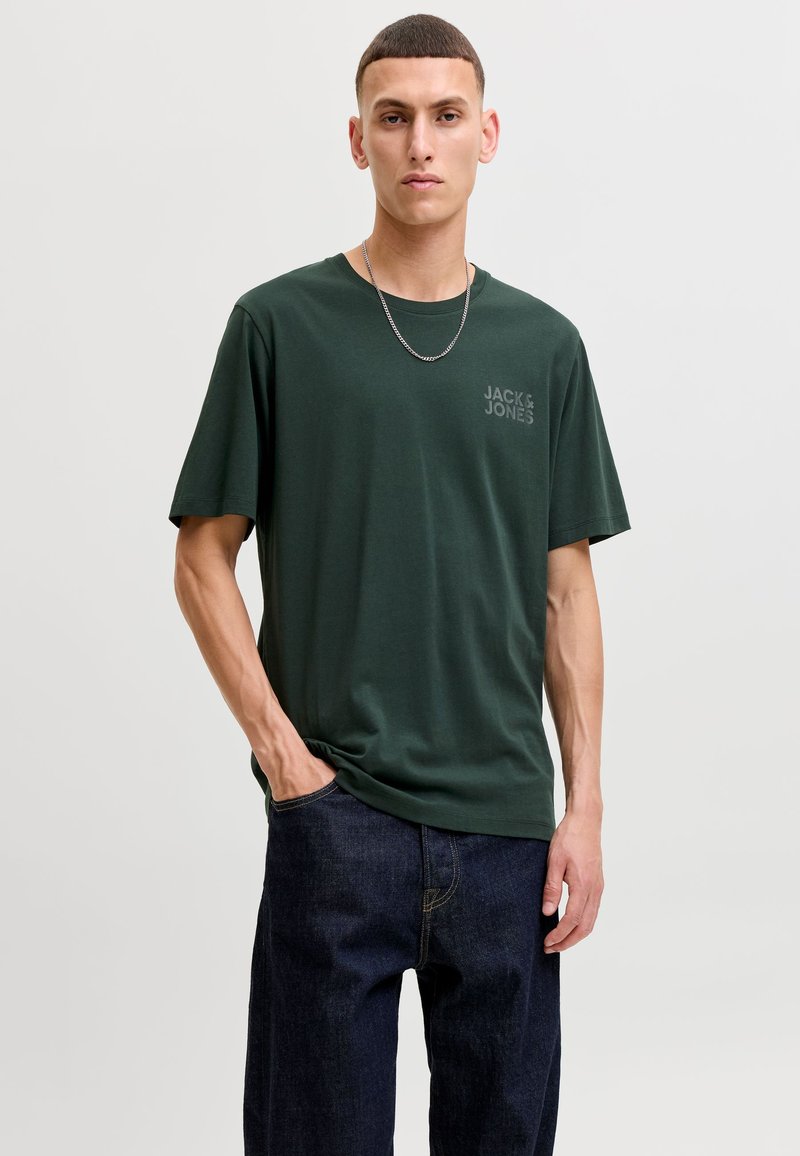 Νέος άνδρας που φοράει σκουροπράσινο T-shirt Jack & Jones, σκούρο τζιν και ασημένιο αλυσίδα λαιμού, στέκεται μπροστά σε ένα απλό φωτεινό φόντο.