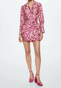 Mujer con un vestido envolvente de manga larga en tonos rosa y blanco con un patrón, de pie contra un fondo liso.