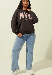Felpa grigia scura con "NYC 1975" in rosa, abbinata a jeans a gamba larga di un blu chiaro e sneakers bianche su uno sfondo neutro.