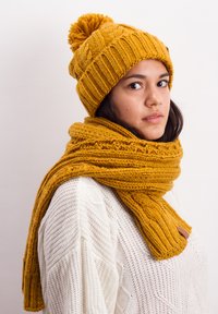 Yellow knit beanie with pom-pom and matching thick scarf, featuring ribbed texture, draped around a white sweater. Soft, warm material.