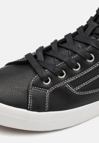 Zapatilla de deporte negra de cuero sintético con punta redonda, suela gruesa de goma blanca, cordones negros, ojales plateados y detalles de costura blanca.