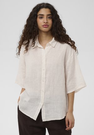 Joven mujer con cabello largo y rizado, vestida con una camisa suelta de color beige claro abotonada y pantalones oscuros, de pie con una mano en el bolsillo contra un fondo liso.