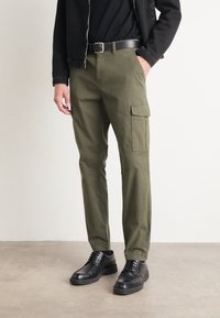 Pantalones cargo verde oliva hechos de algodón, con bolsillos laterales, corte recto y un cinturón. Combinados con zapatos negros con cordones.