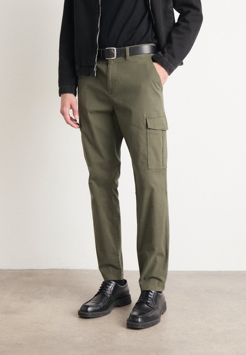 Pantalones cargo verde oliva hechos de algodón, con bolsillos laterales, corte recto y un cinturón. Combinados con zapatos negros con cordones.