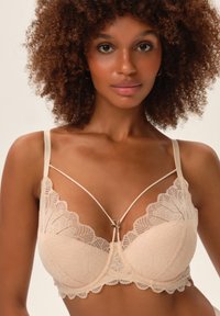 Koronkowy biustonosz w kolorze nude z głębokim wycięciem, falistymi brzegami oraz delikatnym wzornictwem ramiączek z centralnym akcentem w postaci zdobienia.