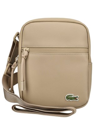 Beżowa torba na ramię z regulowanym paskiem, przednia kieszeń na suwak, teksturowana tkanina oraz małe zielone logo krokodyla w prawym dolnym rogu.