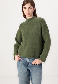 Gebreide trui in donkergroen met een hoge kraag, oversized pasvorm, geribbelde textuur en brede mouwen, gecombineerd met lichtblauwe denim jeans.
