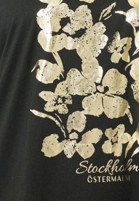 Czarny strój z złotym kwiatowym nadrukiem oraz teksturowymi akcentami. Zawiera napis "Stockholm Östermalm" w złocie.