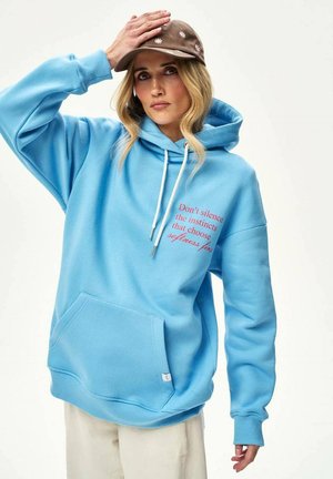 Kobieta w niebieskiej bluzie z kapturem z czerwonym napisem, beżowych spodniach i brązowej czapce z białymi kwiatowymi wzorami, trzymająca czapkę jedną ręką.