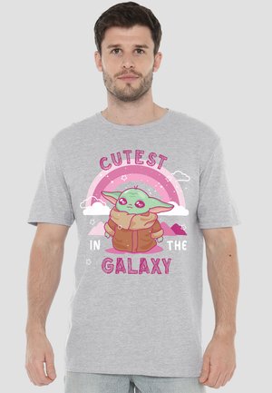 Man wearing gray T-shirt with cartoon green alien in brown coat beneath pink rainbow and text "Cutest in the Galaxy"