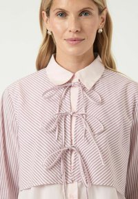 Rosa och vit skjorta med knappar som har ett randigt mönster, klassisk krage och flugdetaljer vid halslinningen. Långa ärmar.