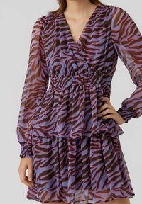 Mujer que lleva un vestido de estampado de cebra en púrpura y marrón oscuro, de manga larga, con escote en V y cintura ceñida, de pie con una mano en la cadera.