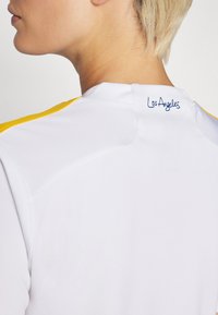 Balts sporta džersijs ar īsām dzeltenām plecu svītrām, uz kura ir izšūts "Los Angeles" zilā krāsā aizmugurē, tuvu kakla izgriezumam.