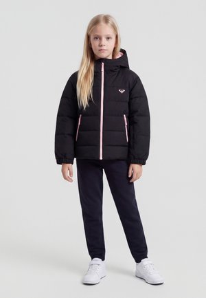 Fille aux longs cheveux blonds portant une doudoune noire avec des fermetures éclair roses, un pantalon foncé et des baskets blanches, debout devant un fond clair uni.