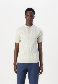 Ljust grå stickad pikétröja med korta ärmar, prydd med en texturerad kabelmönster och treknapps-knäppning, i kombination med blå byxor.