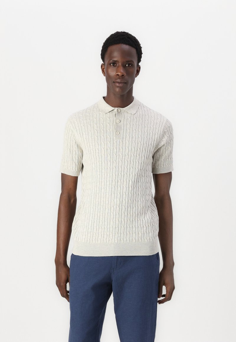 Ljust grå stickad pikétröja med korta ärmar, prydd med en texturerad kabelmönster och treknapps-knäppning, i kombination med blå byxor.