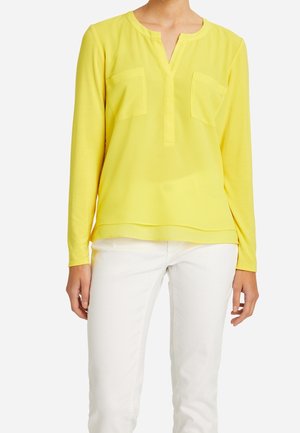 Langærmet gul bluse med V-udskæring, to forlommer og en glat tekstur, kombineret med hvide bukser.