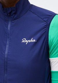 Κοντινό πλάνο ατόμου που φορά μπλε σκούρο γιλέκο με φερμουάρ και λογότυπο "Rapha" πάνω από ποδηλατική φανέλα με μακριά μανίκια σε πράσινο και λευκό.