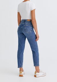 Jeans de denim azul de tiro alto con un corte ceñido, que cuentan con dos bolsillos traseros y un parche de cuero en la cinturilla, combinados con zapatillas blancas.