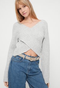 Szary dzianinowy sweter z dekoltem w serek i krzyżującym się wzorem, zestawiony z niebieskimi dżinsami i jasnobeżowym paskiem z okrągłą klamrą.