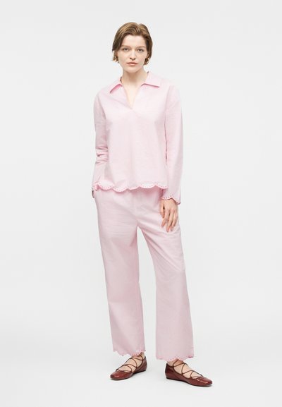 Νεαρή γυναίκα στέκεται φορώντας ελαφρύ ροζ μπλούζα με κυματιστές άκρες και παντελόνι, μαζί με καφέ δερμάτινα παπούτσια με κορδόνια, σε απλό λευκό φόντο.