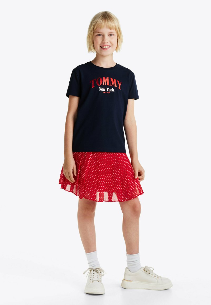 T-shirt navy con la scritta "TOMMY New York", abbinato a una gonna rossa a pois. Sneakers bianche e calzini bianchi a costine completano l'outfit.