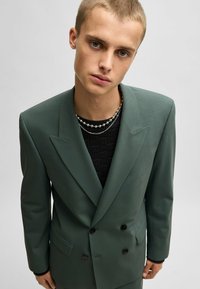 Doppelreihige Blazer aus dunkelgrünem Stoff, mit scharfen Revers, zwei Fronttaschen und schwarzen Knöpfen. Über einem strukturierten schwarzen Pullover getragen.