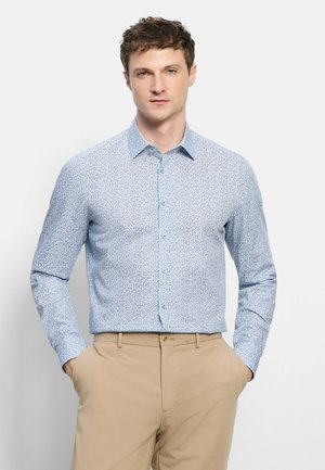 Homme portant une chemise à manches longues bleu clair à motifs et un pantalon beige, debout les mains dans les poches devant un fond uni.