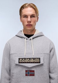 Grå sweatshirt med en frontlomme, der har et logo patch i sort og beige, samt et lille patch af det norske flag i rødt og blåt. Blødt stof, med hætte.