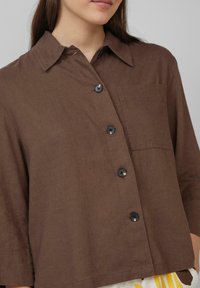 Bruine linnen blouse met een kraag, voorzien van een patchzak, zwarte knopen en een ontspannen pasvorm, die een gladde, gestructureerde uitstraling biedt.