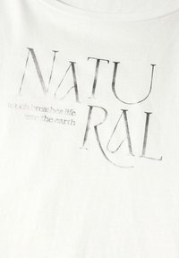 Camiseta de algodón blanca con texto metálico plateado que dice "NATURAL" y una frase más pequeña debajo en una fuente sencilla.