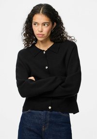 Črni pleteni pulover z ovratnikom, z zlatim gumbom v obliki cvetja in rahlo skrajšanim robom, nošen čez modri denim.