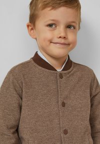 Brązowa kurtka w jodełkę z brązowym kołnierzem, zapinana z przodu na guziki, z bocznymi kieszeniami. Miękka w dotyku, o dopasowanym kroju.