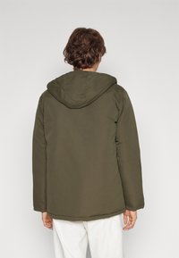 Persona con pelo corto castaño, vestida con una chaqueta verde con capucha y pantalones blancos, vista desde atrás contra un fondo liso.