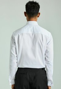 Cămașă albă cu textură netedă, guler în vârf și mâneci lungi, având mansete accentuate și croială ajustată.