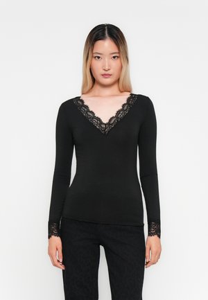 Kvinde med glat ombre-hår iført en tætsiddende sort langærmet top med blondedetaljer på V-udskæringen og ærmekanter, stående mod en hvid baggrund.