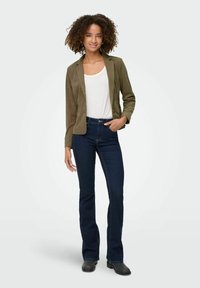 Olivgrüner Cordblazer mit taillierter Passform, kombiniert mit einem weißen Tanktop und dunkelblauen Schlagjeans. Schwarze Stiefeletten, entspannte Pose.